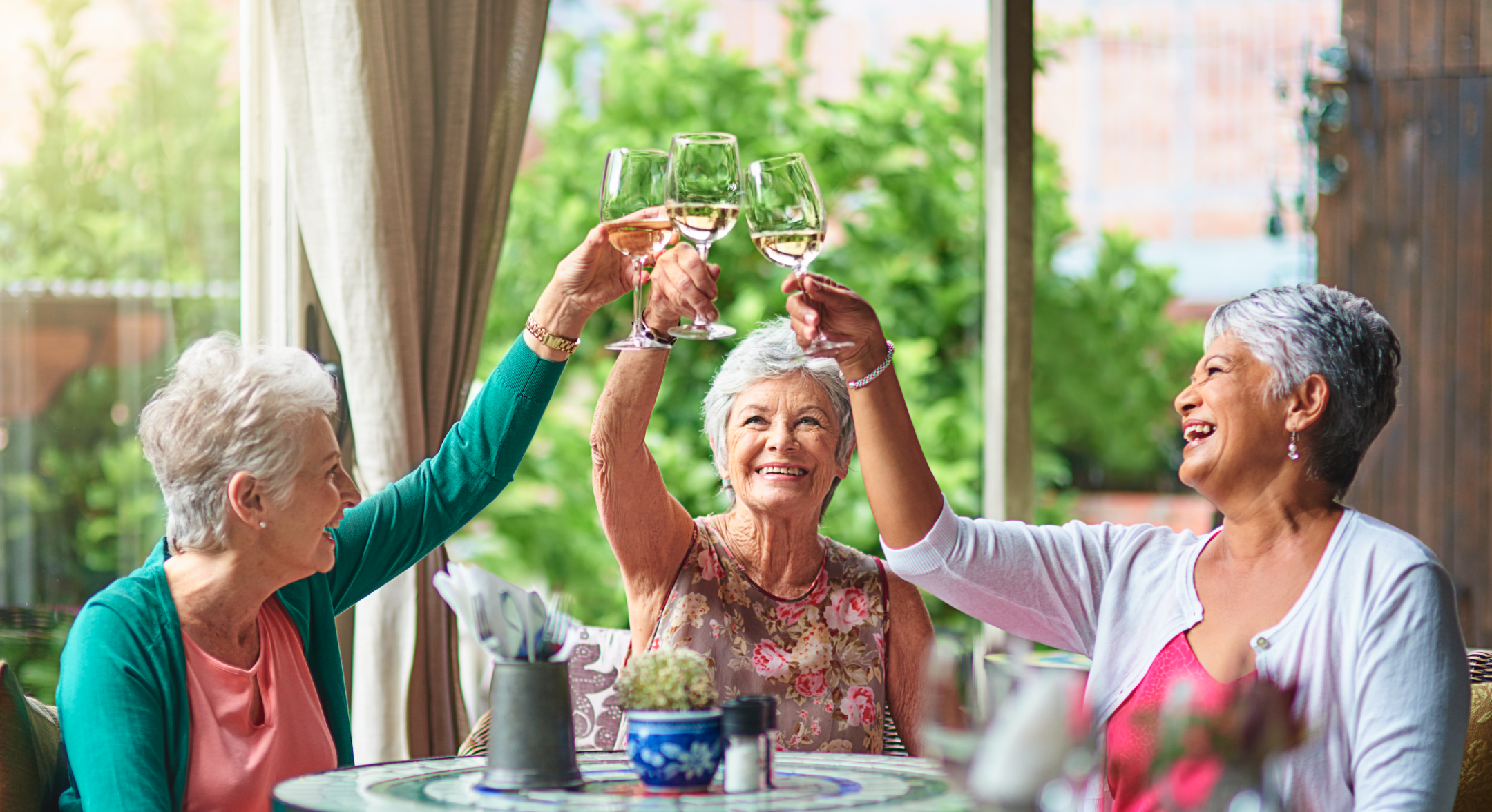 Three smiling older women sit at a table outdoors, raising wine glasses in a cheerful toast. The background is green and sunny, creating a warm, joyful atmosphere.